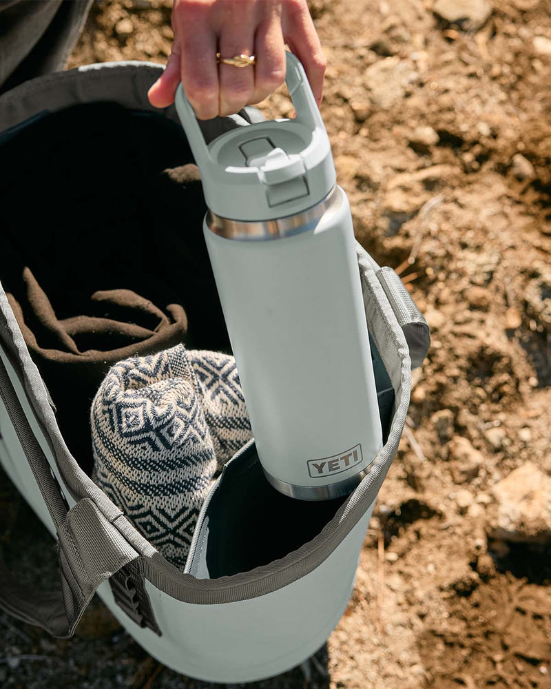 A straw cap water bottle being placed inot a YETI tote bag outdoors.