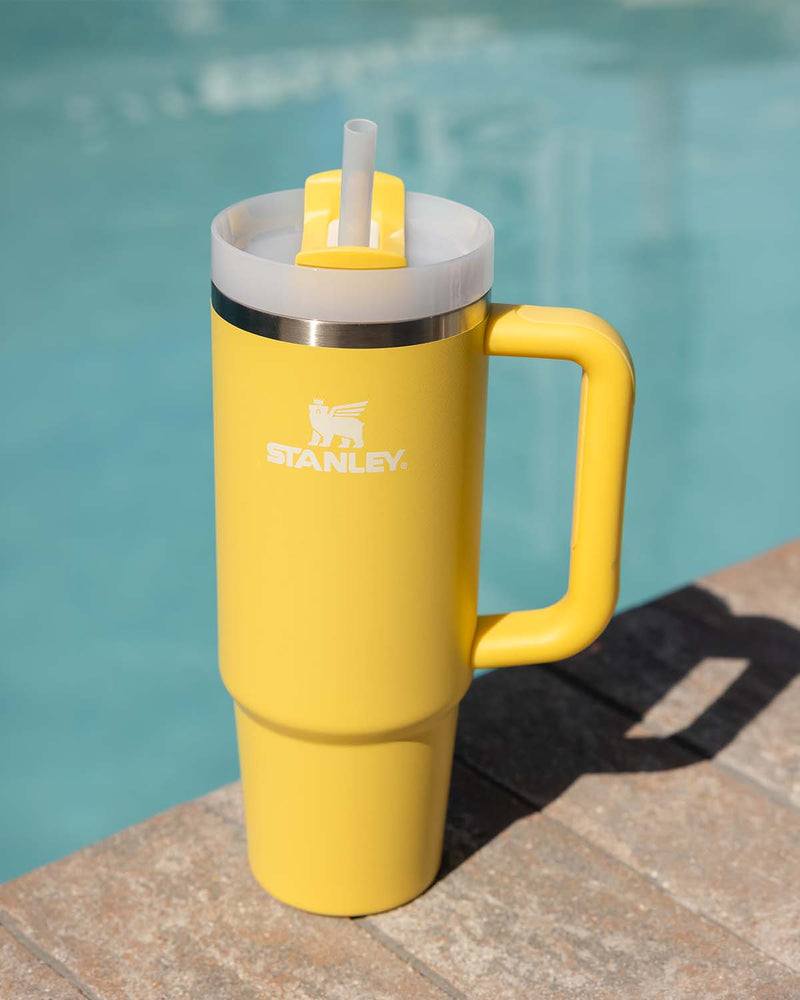 Yellow Stanley handled tumbler with straw lid placed on poolside stone ledge in bright sunlight.