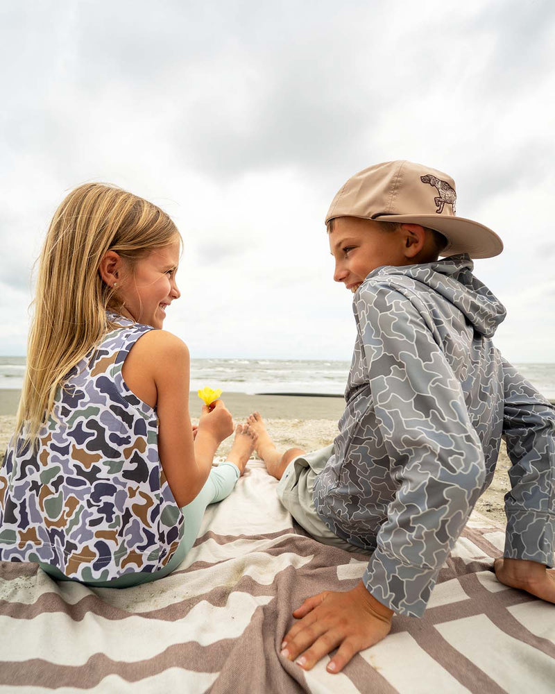 Two kids sitting on a towel on the beach in camo shirts from Local Boy and Local Girl.