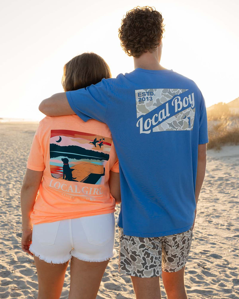 Local Boy and Local Girl graphic t-shirts worn by couple standing on beach at sunset, shown from back.