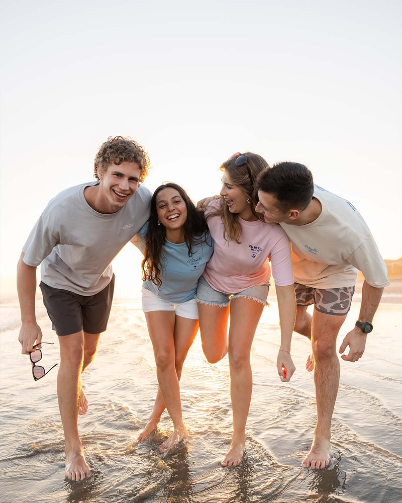 Group of four friends wearing casual t-shirts and shorts walking barefoot along shoreline at sunset.