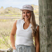 Woman wearing a white sleeveless top and light-colored cap with a fish design, standing outdoors.