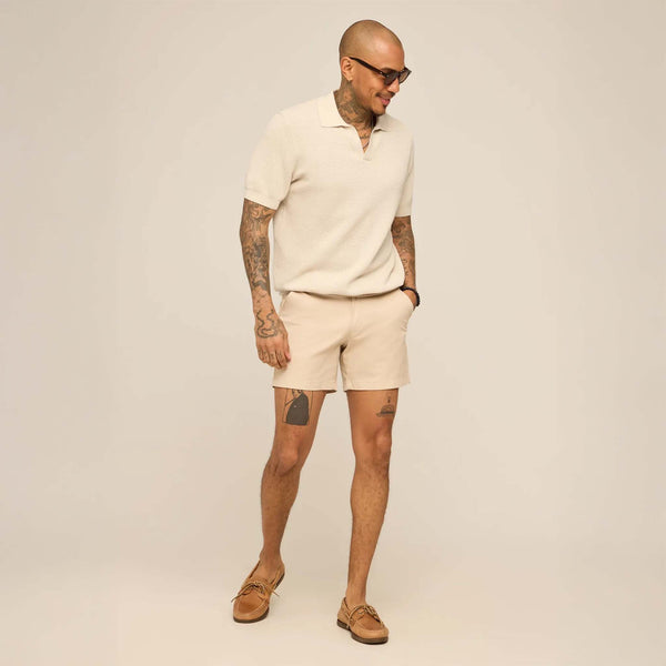 Man wearing a beige polo shirt and shorts with sunglasses on a plain background