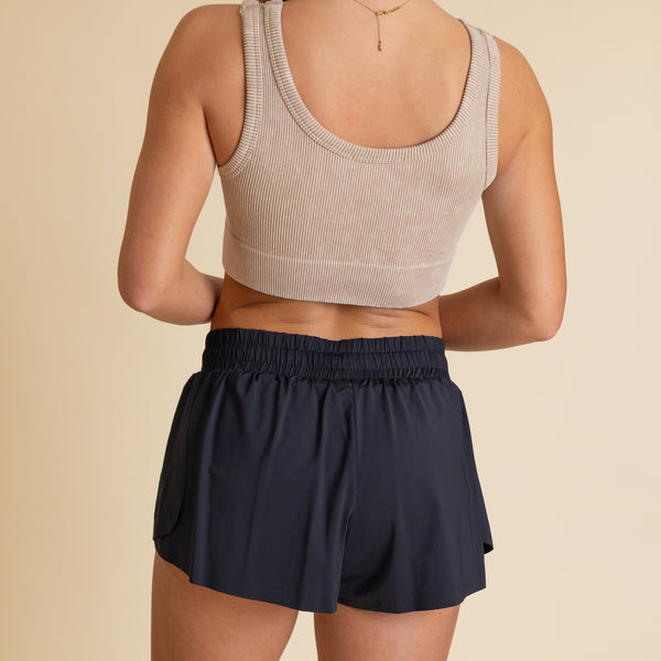  Back of woman wearing a beige brami tank and black elastic waistband athletic shorts.