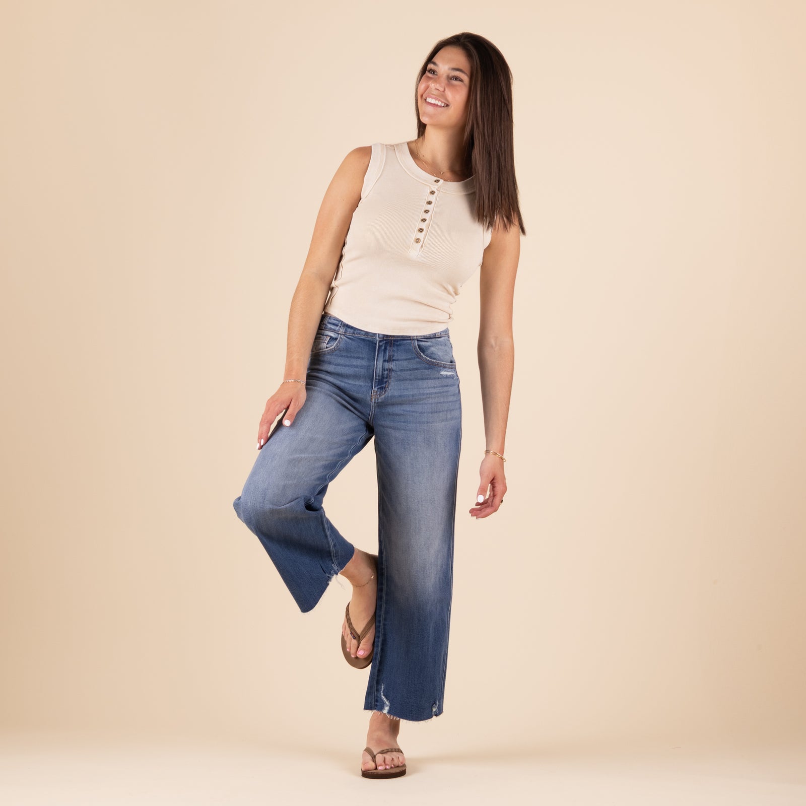 Front view of a woman wearing high-rise, medium-wash wide leg jeans with a faded front, subtle whiskering, light distressing near the hem, and raw edge details. She is also wearing a sleeveless beige top and sandals, standing against a neutral background.