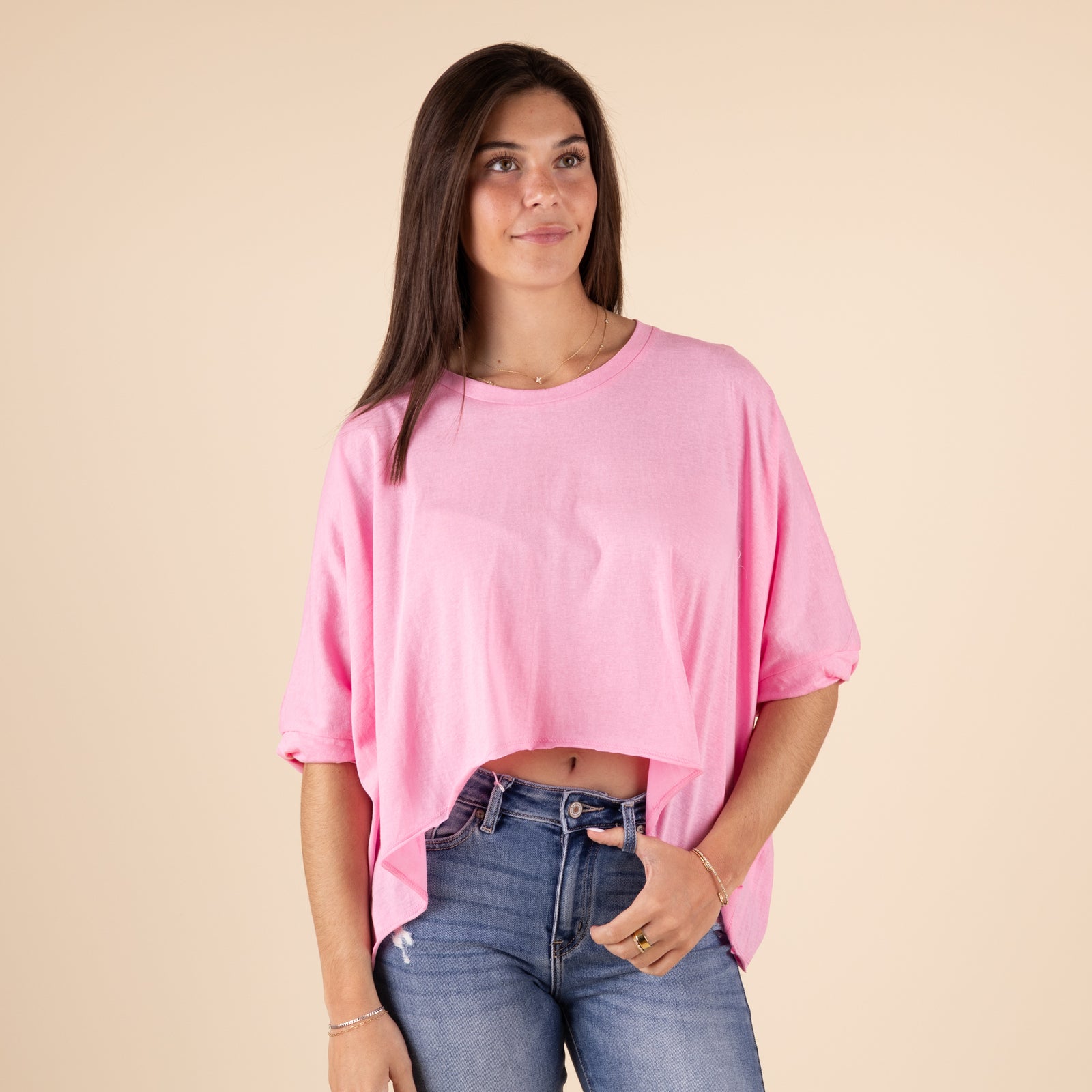 Women's Double Zero Hi Lo Twist Short Sleeve Top, color pink, sizes S-L.
A woman wearing a bubblegum pink oversized short-sleeve crop top with a wide, asymmetrical hemline that forms a pointed shape in the front. The shirt has rolled sleeves and is paired with light-wash high-waisted jeans. The background is a neutral beige color.