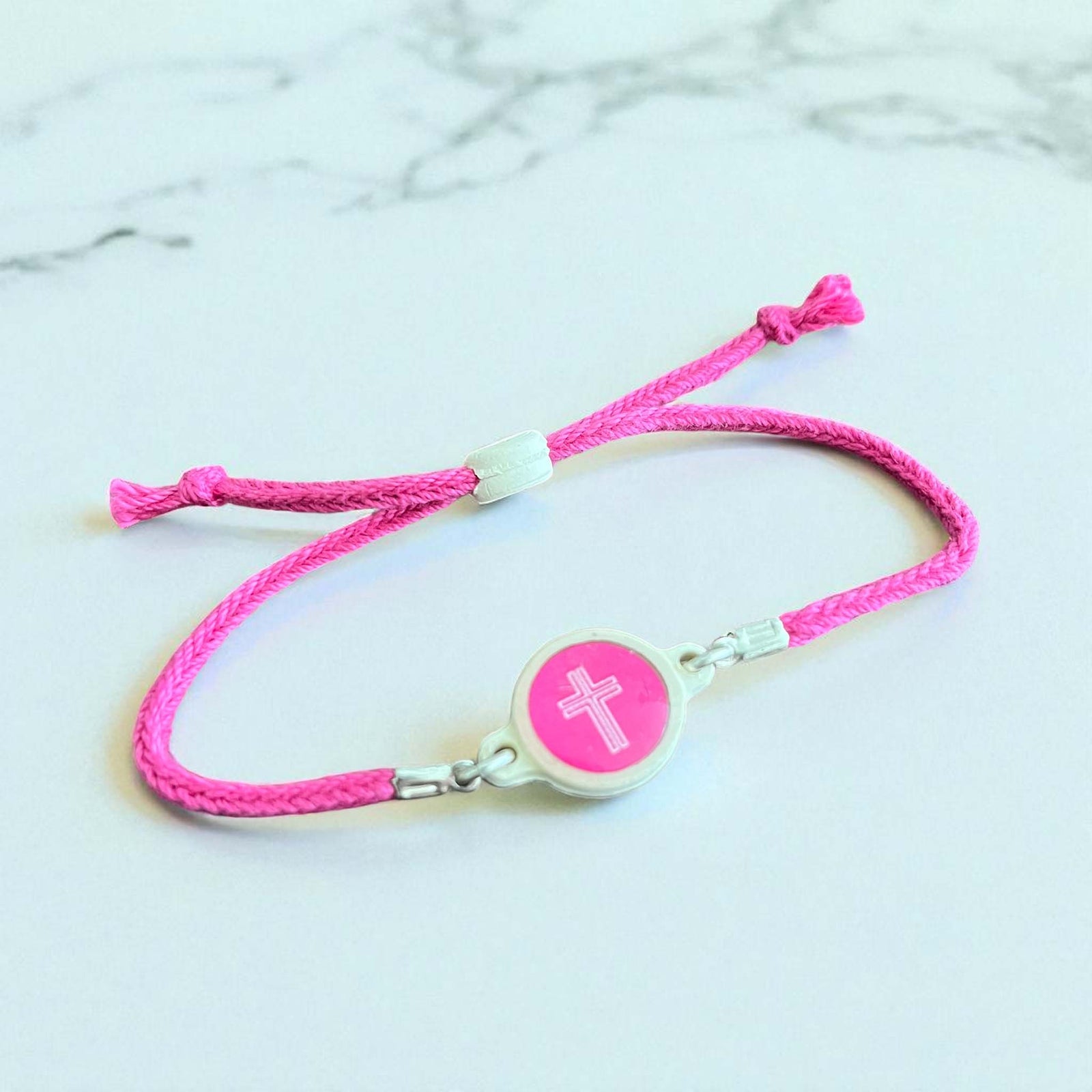 Pink braided bracelet with adjustable ties, silver accents, and round pink charm with white cross, shown on white background.