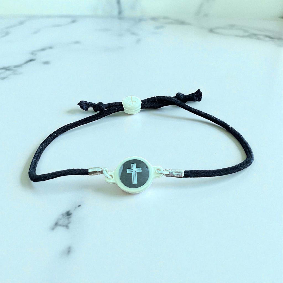 Black cord bracelet with a silver clasp and cross charm on a marble background