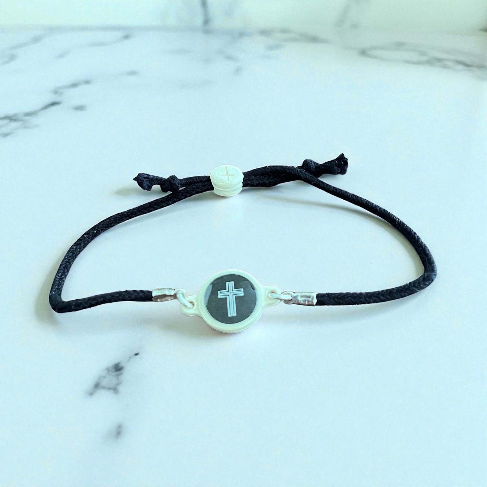 Black cord bracelet with a silver clasp and cross charm on a marble background