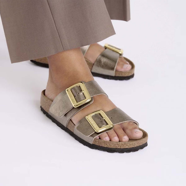 Birkenstock Sydney Luxe Buckle Sandals in Taupe, sizes 5/36, 6/37, 7/38, 8/39, 9/40, 10/41, 11/42. Model wearing sandals with brown pants; close-up highlights foot positioning and shiny buckles.
