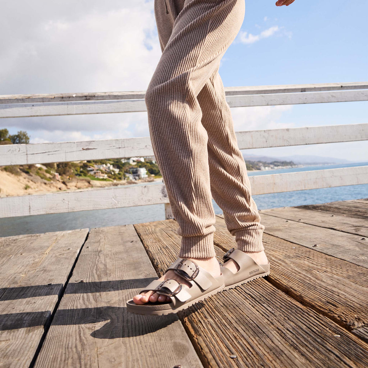 Person wearing beige sandals and pants standing on a wooden dock with a scenic background.