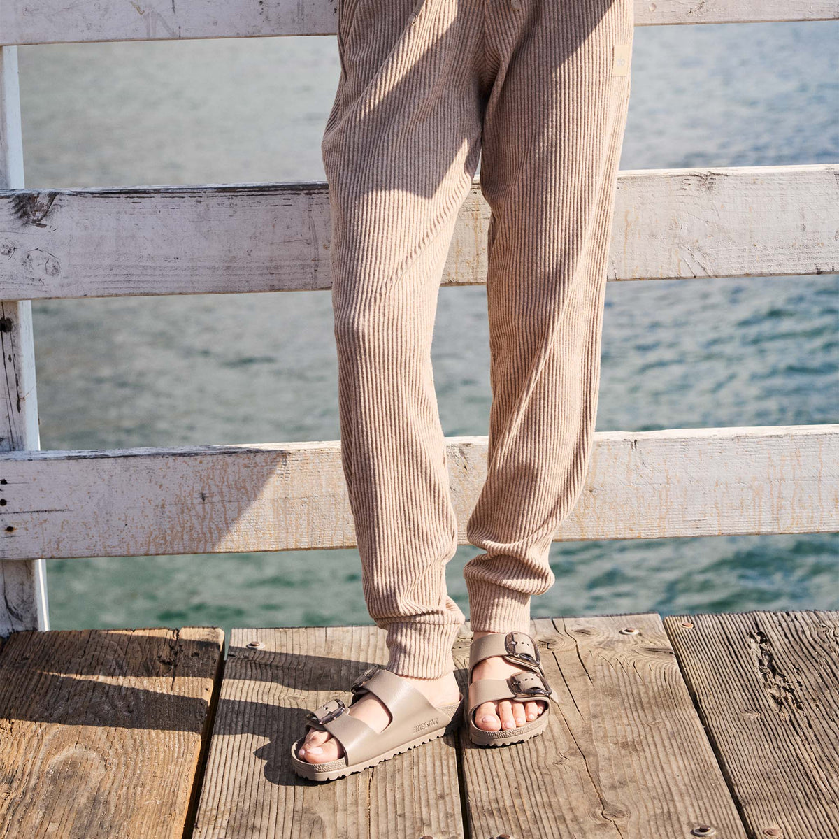 Person wearing beige pants and sandals standing on a wooden dock with water in the background