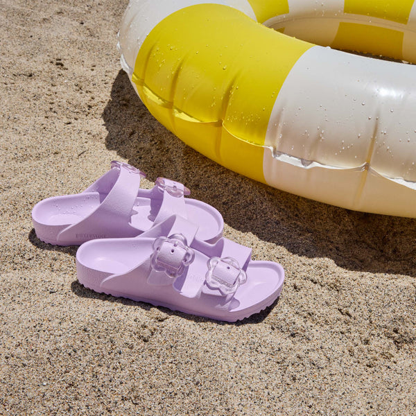 Birkenstock Youth Purple sandal with two flower buckles shown on the sand next to a water toy.