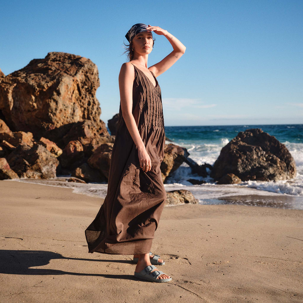 Lifestyle image of a model standing on a beach wearing a flowing brown dress and Pure Sage Birkenstock Arizona EVA sandals with translucent buckles and two adjustable straps.