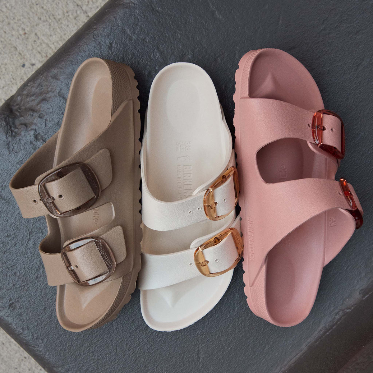 Three pairs of Birkenstock Arizona EVA sandals in neutral colors including Pink Clay, cream, and taupe with translucent buckles displayed on a dark surface.