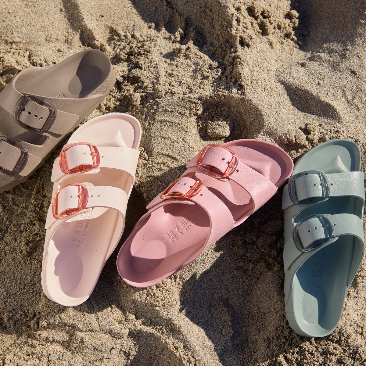 Multiple Birkenstock Arizona EVA sandals in Pink Clay, cream, taupe, and sage arranged on beach sand with translucent buckle straps.
