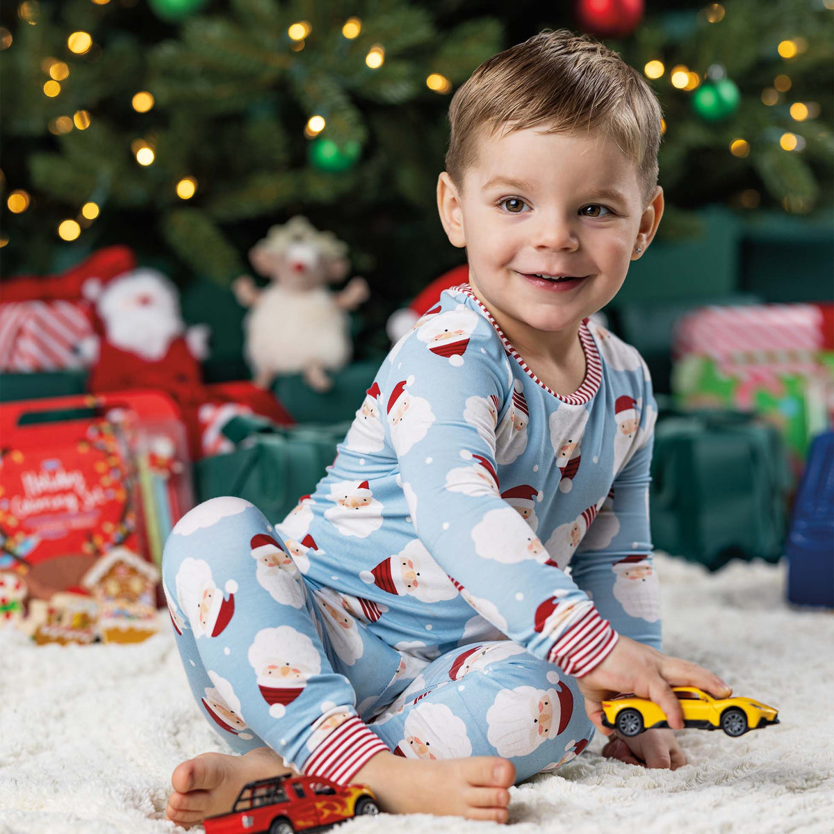 toddler boy in front of a christmas tree wearing mud pie Toddler Santa Glow Pajamas in Blue.