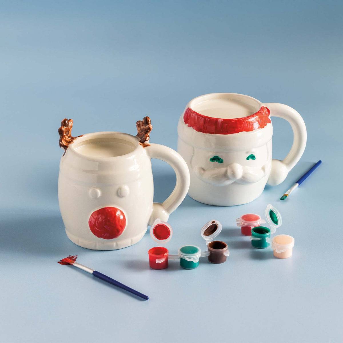 Painted ceramic DIY mugs in reindeer and Santa designs with paintbrushes and open paint pots displayed on a light blue background.