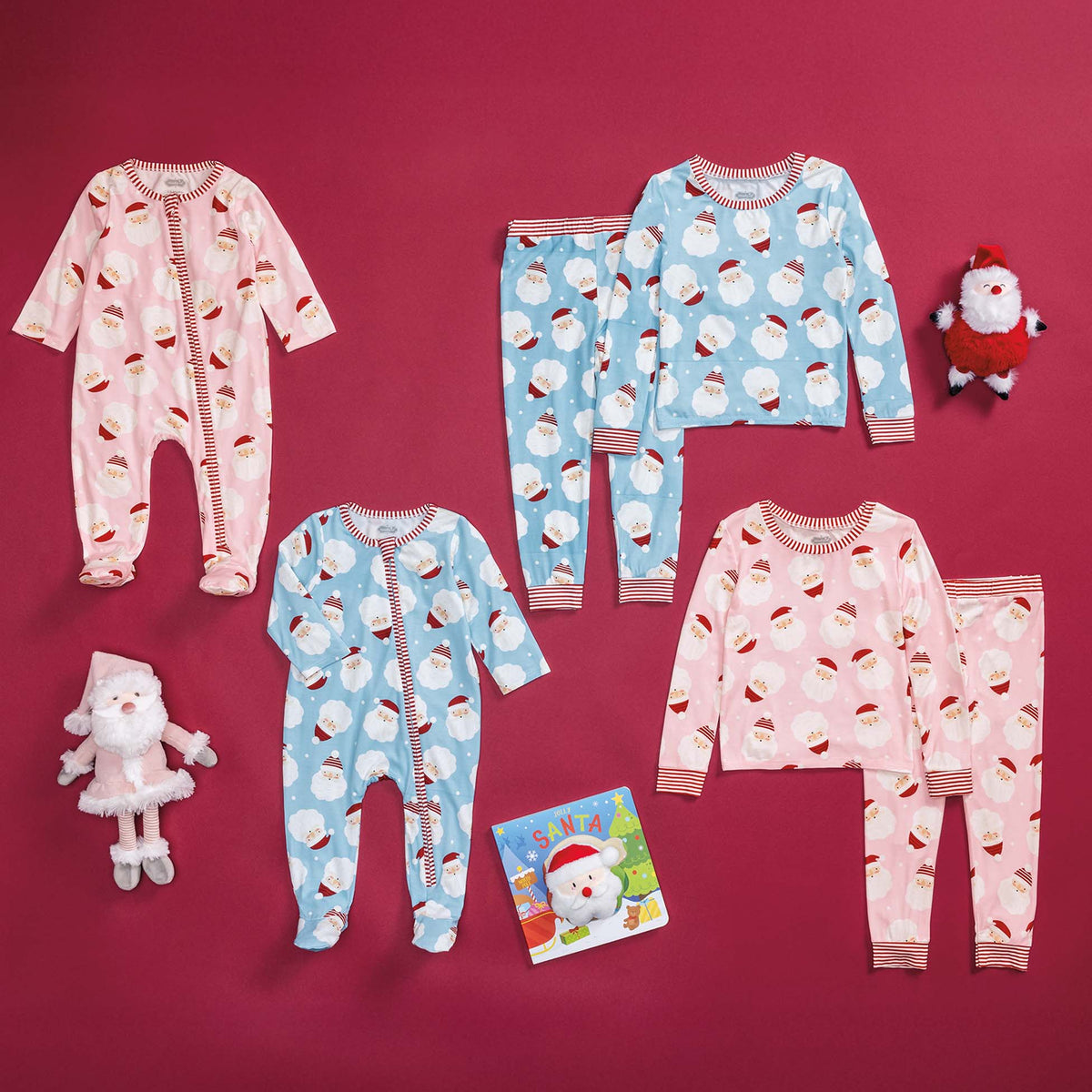 Flat lay of toddler and infant pajamas in pink and blue Santa face prints, including sleepers and two-piece sets, styled with plush toys and a holiday book.
