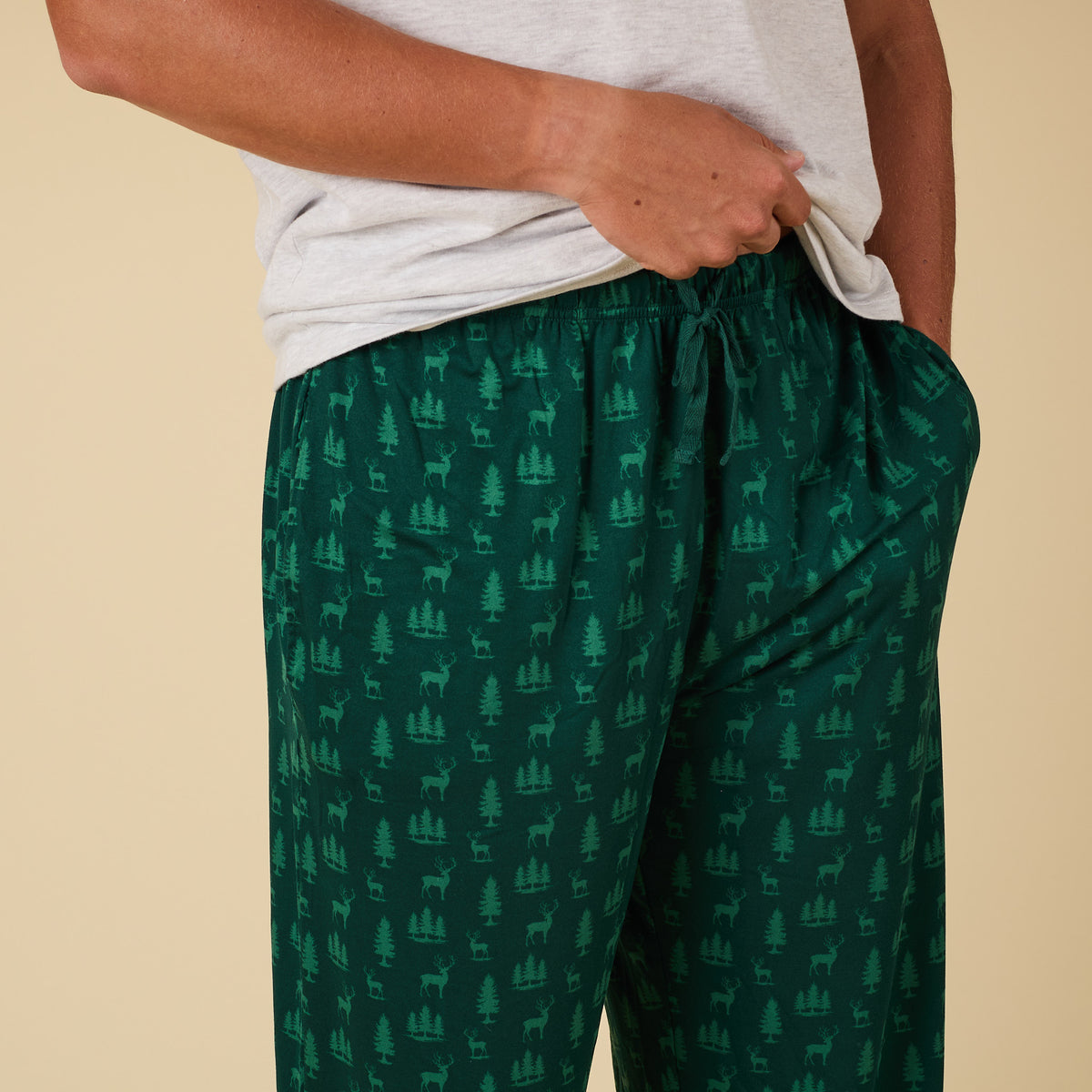 Green pajama pants with deer and tree pattern worn by a person on a beige background