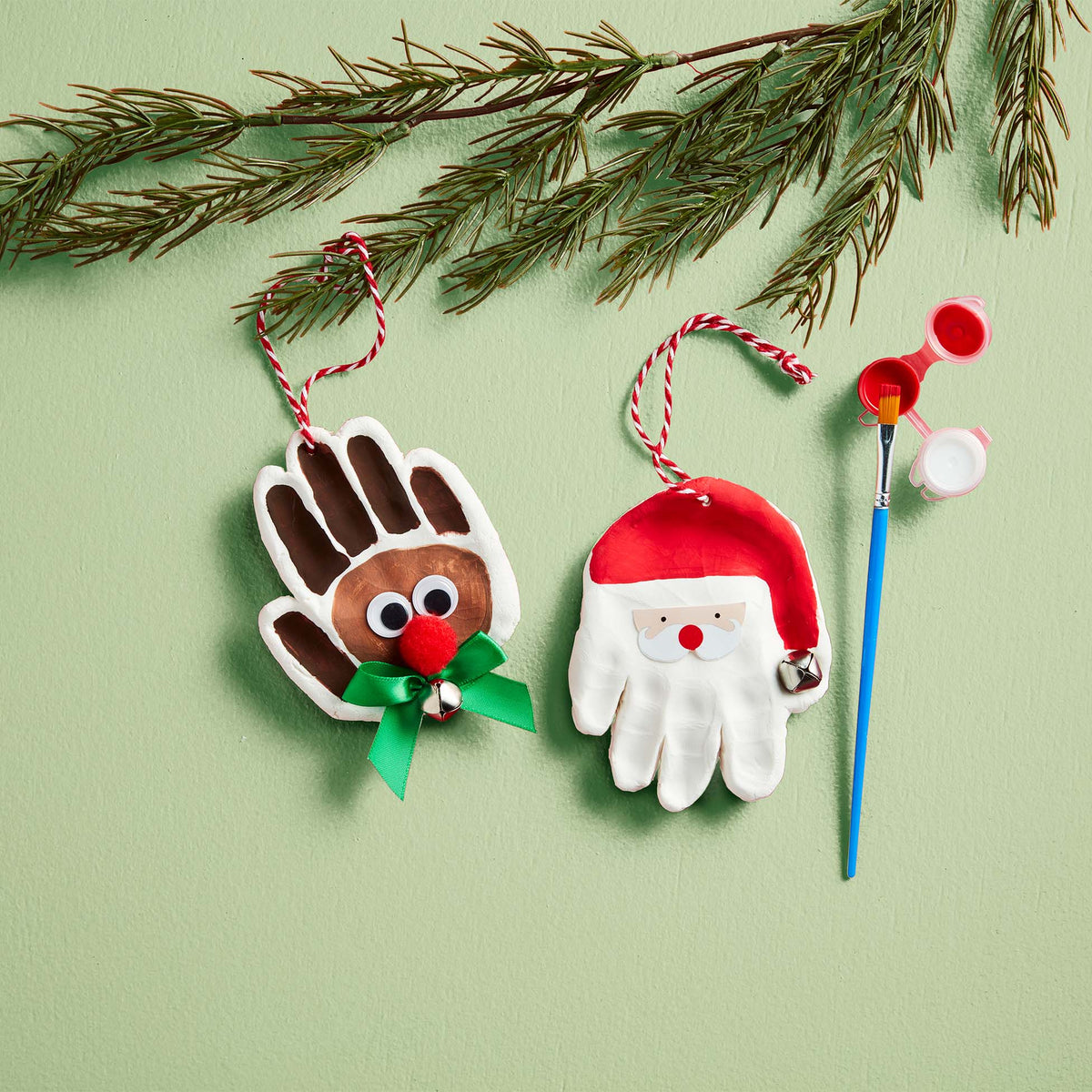 Painted handprint ornaments shaped like a reindeer with a red nose and Santa with a hat, displayed with paintbrush and paints.
