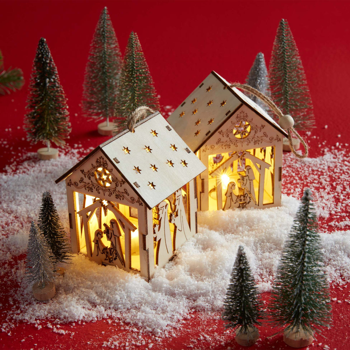 Two wooden light-up nativity house ornaments glow warmly in faux snow, surrounded by mini-Christmas trees on a red background.