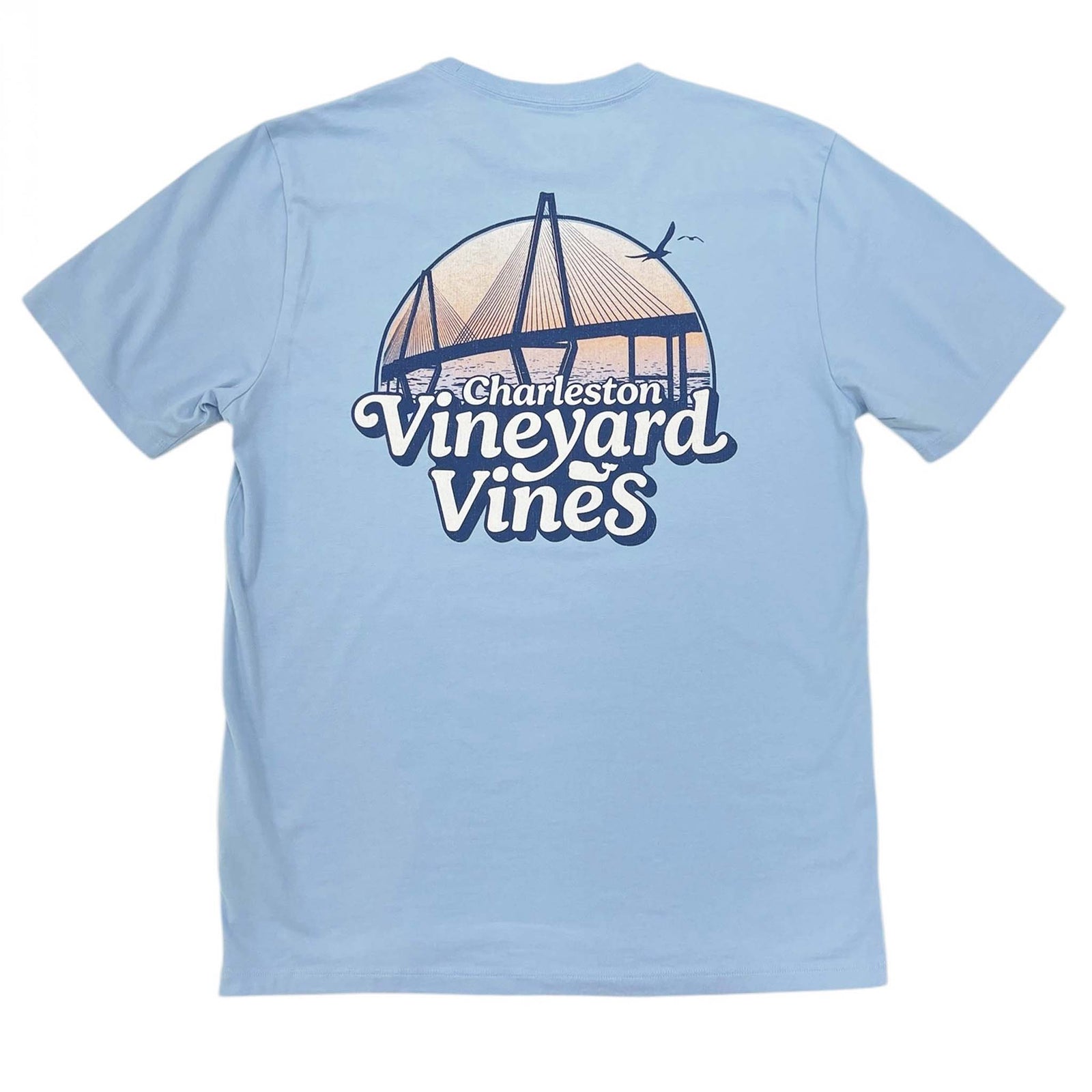 Vineyard Vines Charleston Bridge Short Sleeve T-shirt, color light blue, sizes S-XXL, 100% cotton.  Back of a light blue Vineyard Vines short sleeve t-shirt featuring a blue sketch-style illustration of Charleston’s iconic bridge. A sunset gradient and harbor water create a colorful backdrop behind the bridge. Below the design, 'Vineyard Vines' and 'Charleston' are written in a retro-style font.