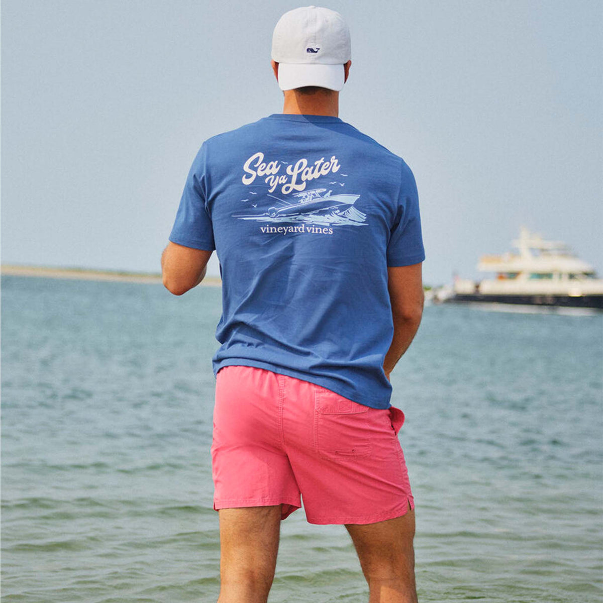 Person wearing a blue t-shirt with a graphic and text, pink shorts, and a white cap, standing by the water.