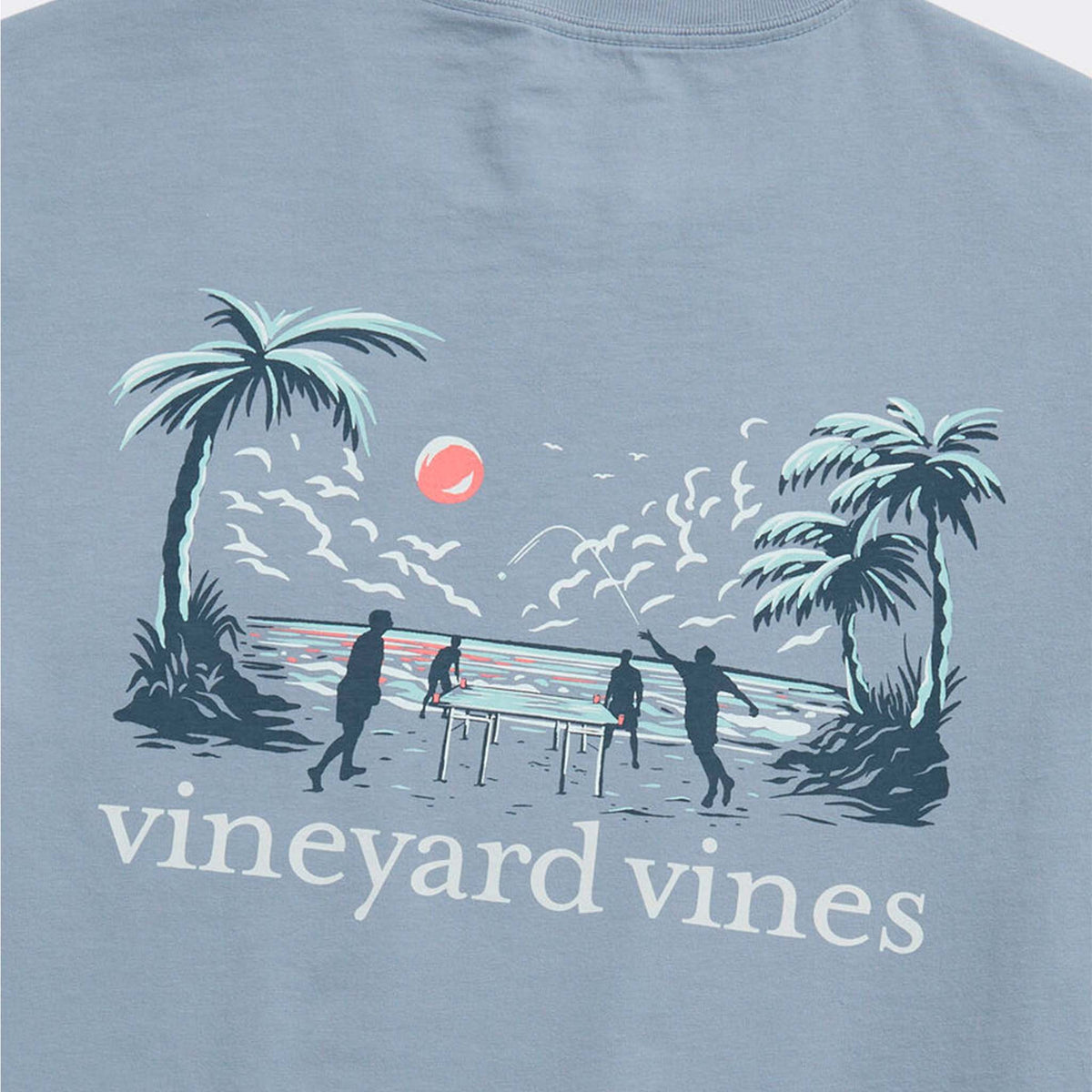 Close-up of back graphic showing palm trees, ocean waves, red sun, and silhouettes playing beach game above Vineyard Vines logo.