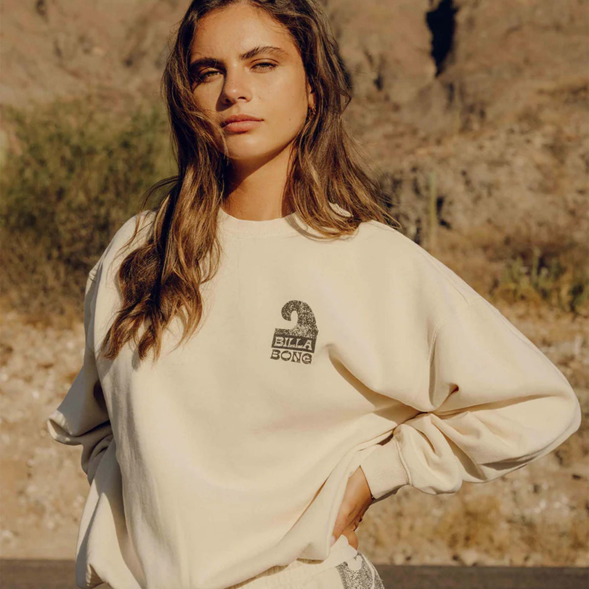 Outdoor view of woman wearing cream Billabong sweatshirt with wave logo and relaxed fit.