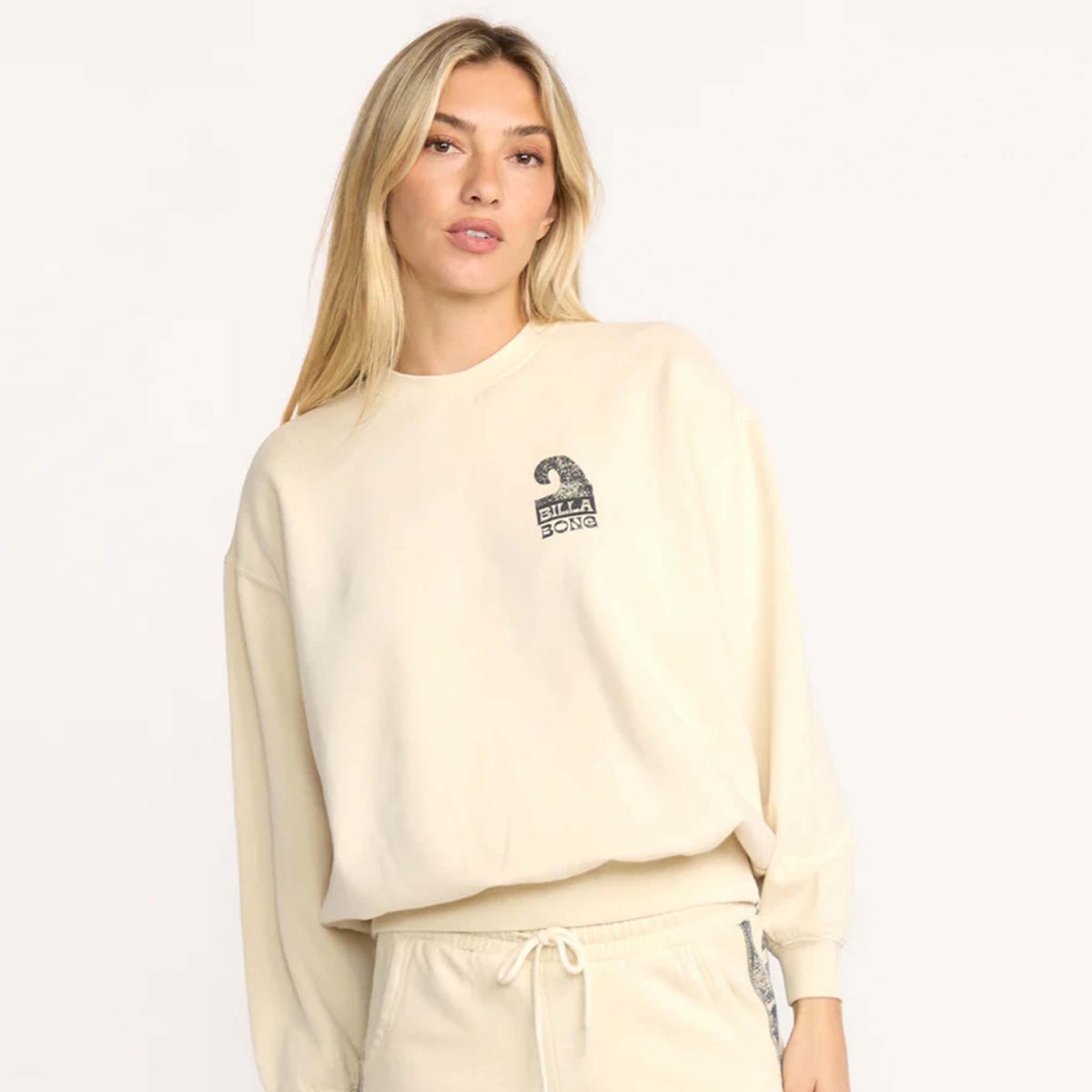 Woman wearing a cream Billabong sweatshirt with small wave logo graphic on the front.