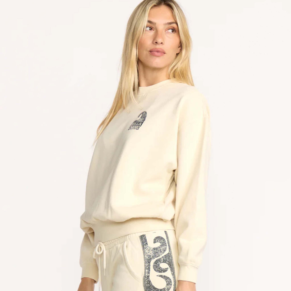 side view of Woman wearing a cream Billabong sweatshirt with small wave logo graphic on the front.