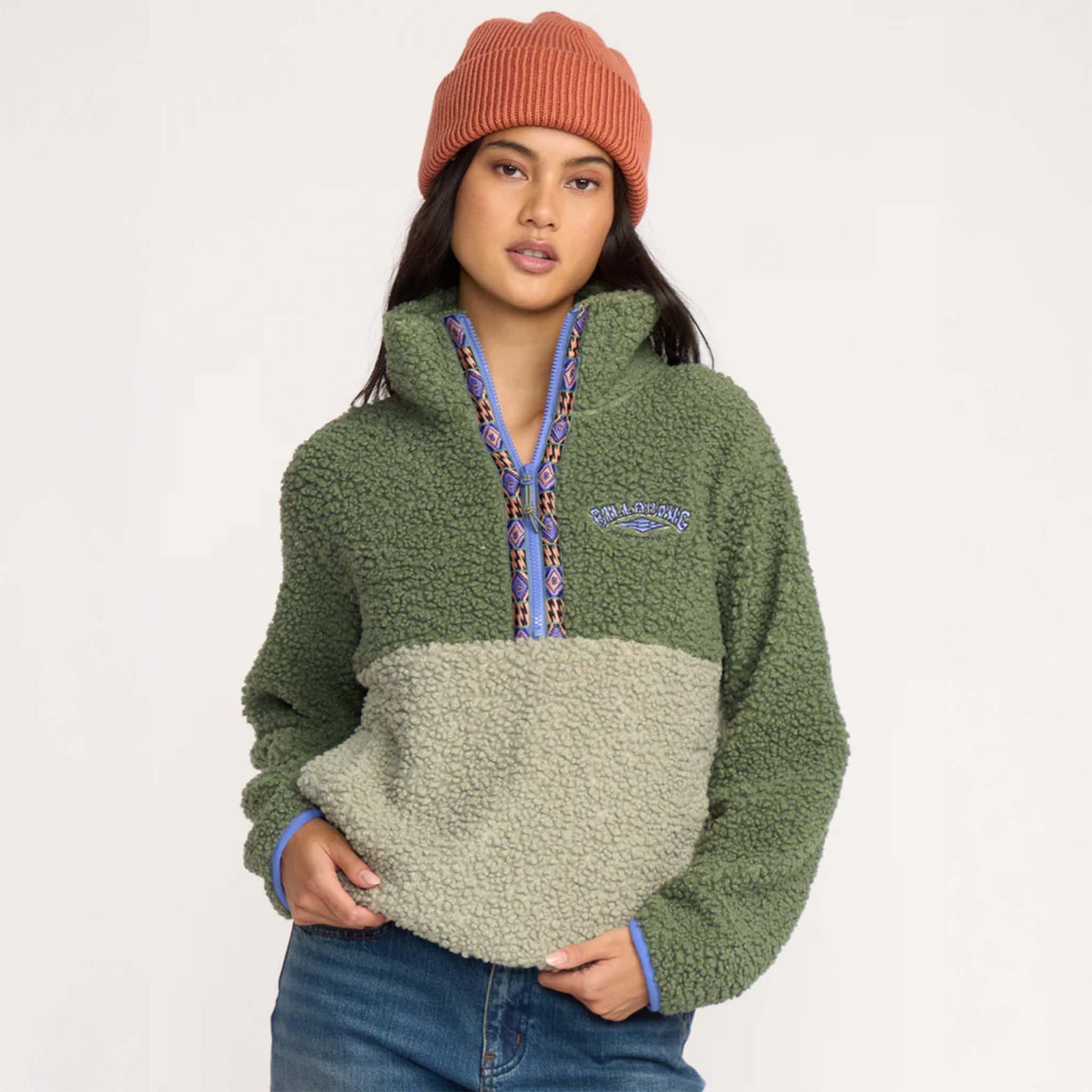 Woman wearing a two-tone green sherpa pullover with embroidered Billabong logo and patterned half-zip detail, paired with jeans and an orange beanie.