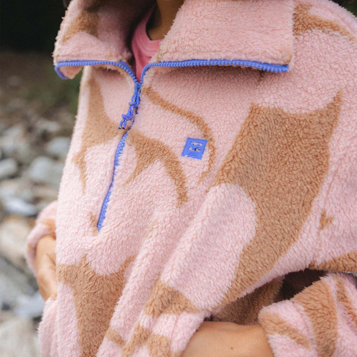 Close-up of pink and tan sherpa pullover showing blue zipper and embroidered logo patch.