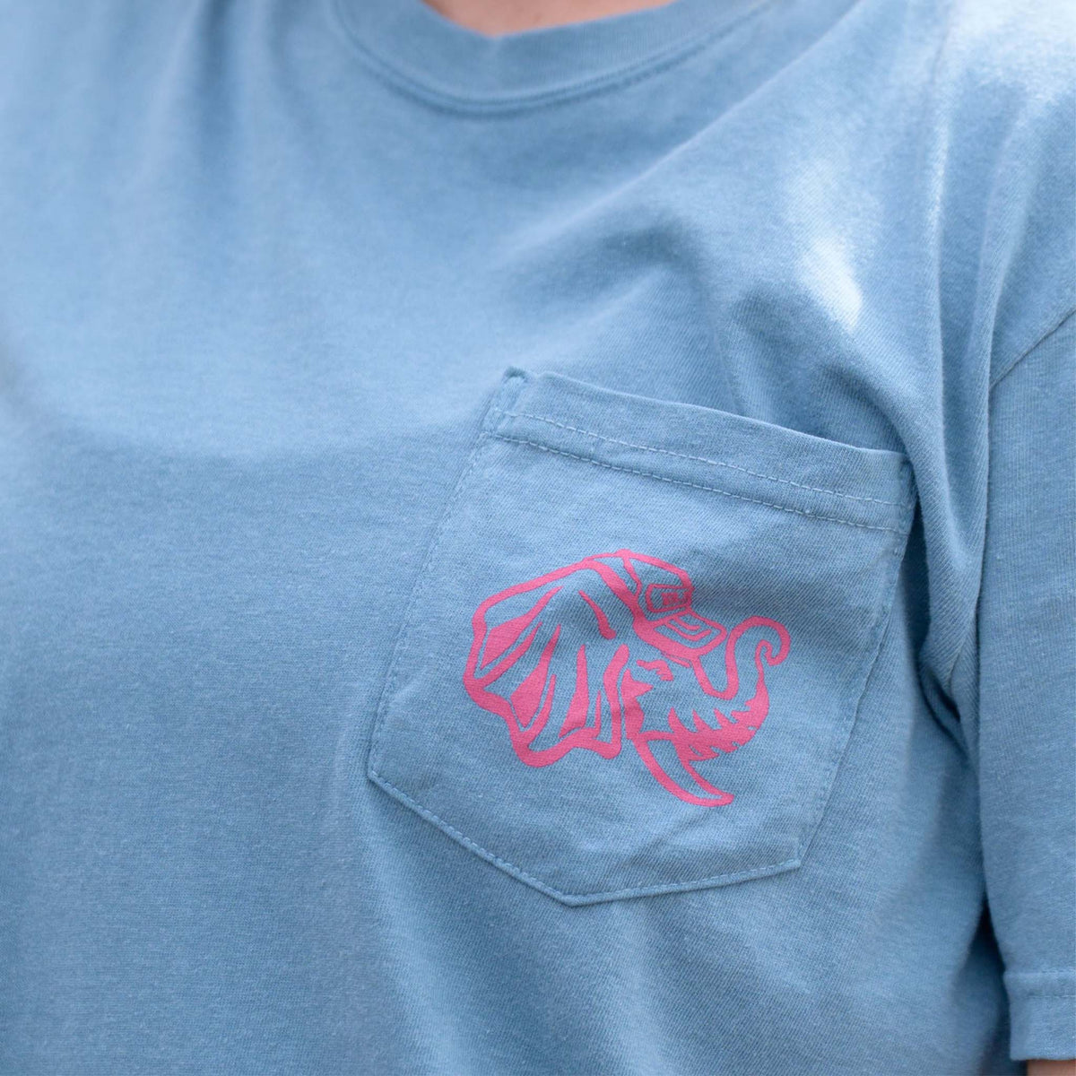 Close-up of Big Al Grounds Crew pocket tee in ice blue with pink elephant logo on pocket.