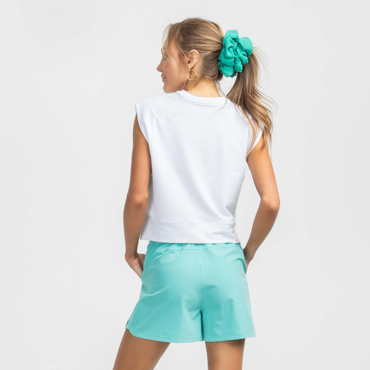 Woman wearing a white sleeveless top and turquoise shorts on a plain background