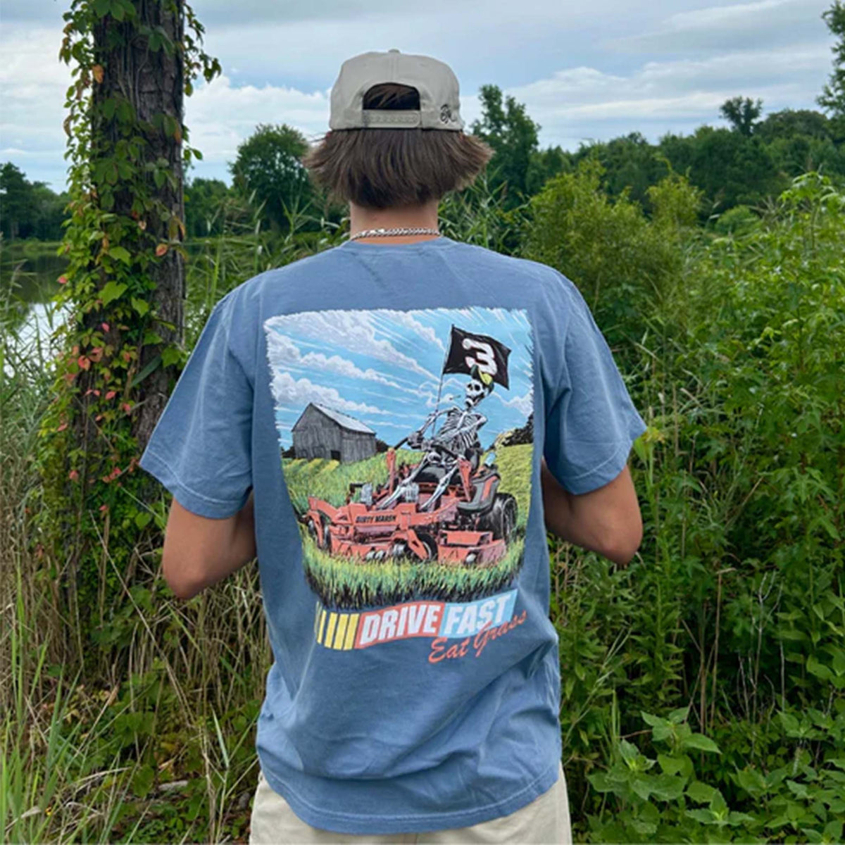 Person wearing a blue Dirty Marsh t-shirt with a large back graphic of a skeleton mowing grass with a “Drive Fast Eat Grass” slogan.