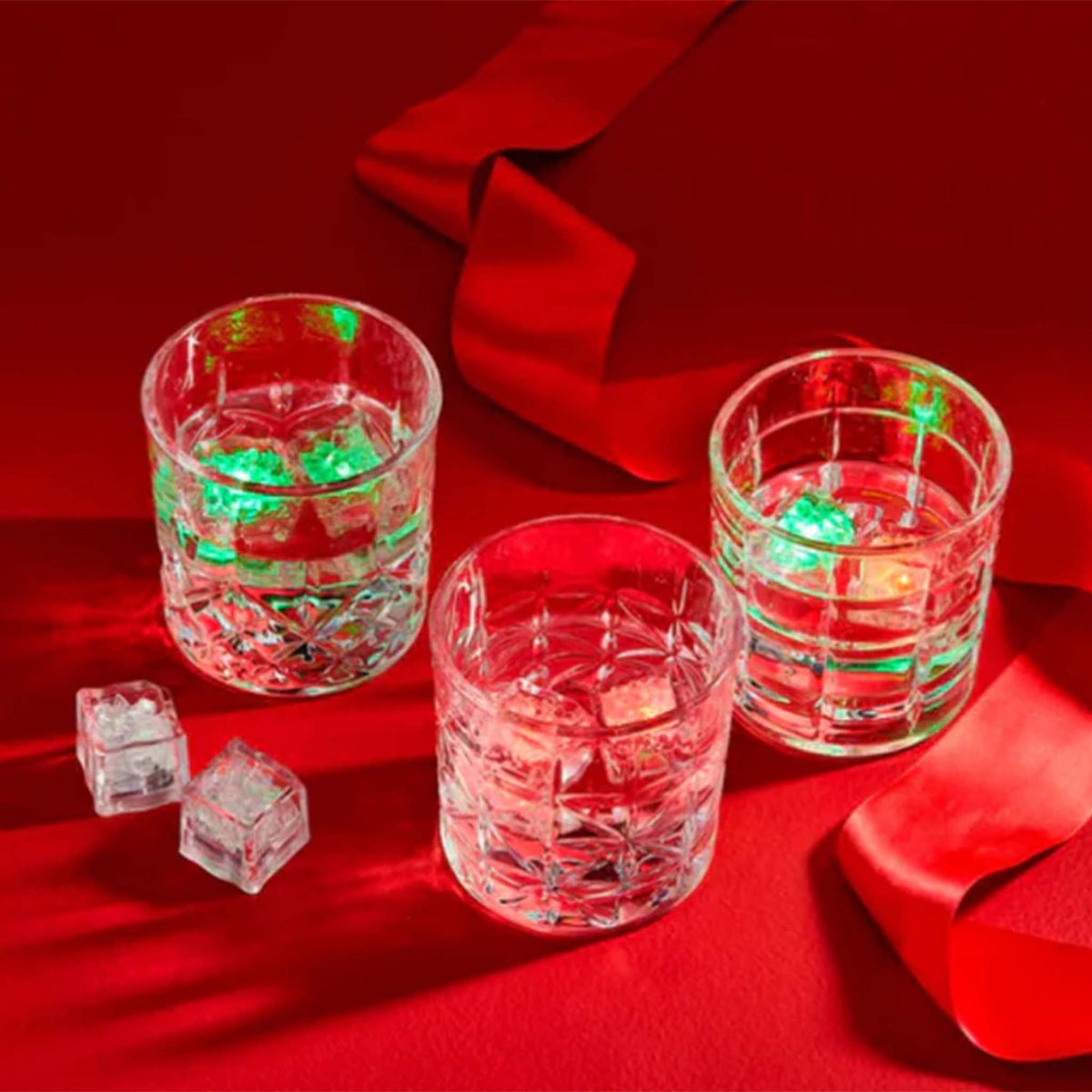 Three clear glasses on a red background with glowing red and green light-up drink cubes inside, creating a festive holiday effect.