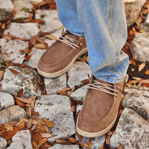 Brown suede shoes worn with blue jeans on a stone path with fallen leaves.