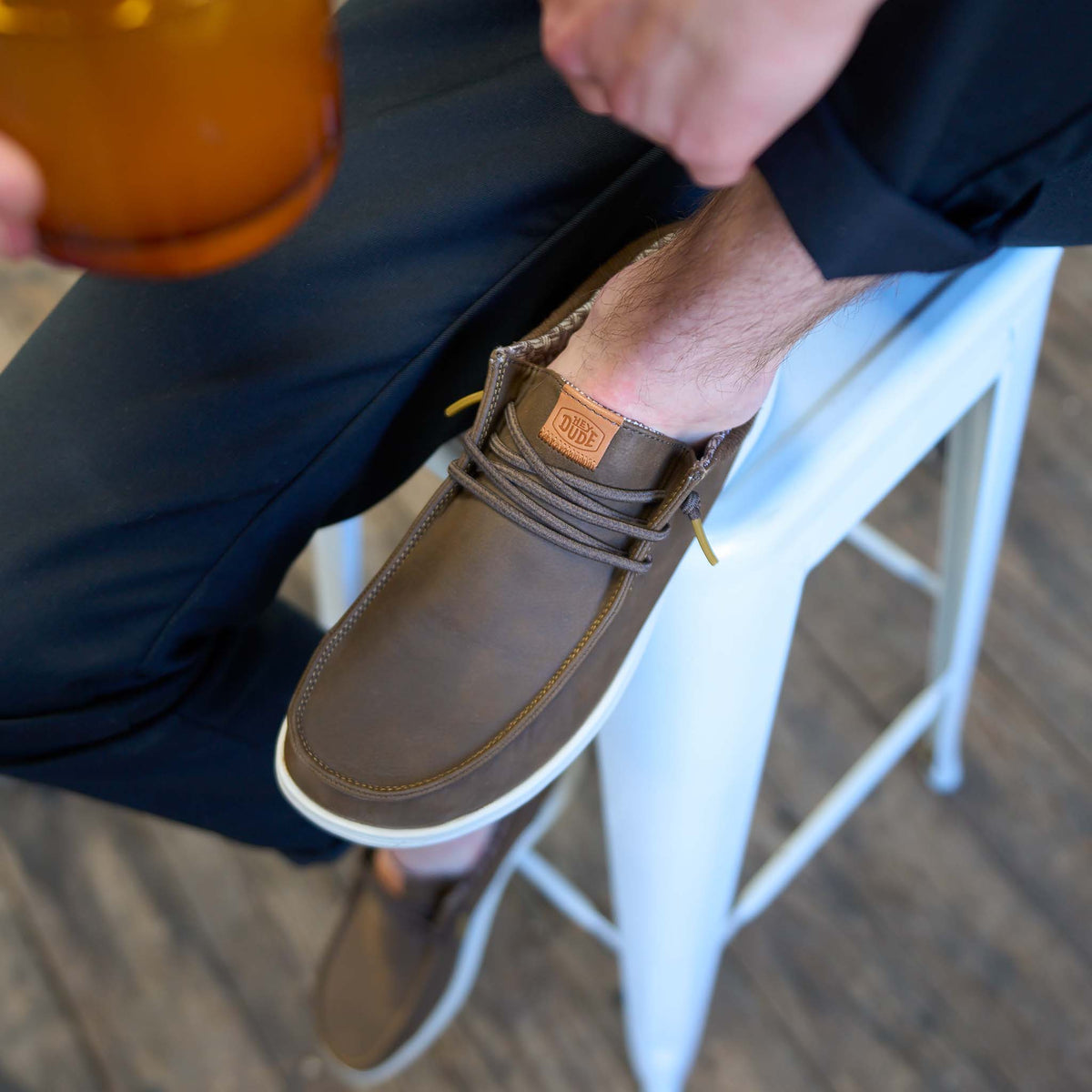 Lifestyle shot of Hey Dude Paul Classic shoe in brown worn with dark pants, shown resting on stool.