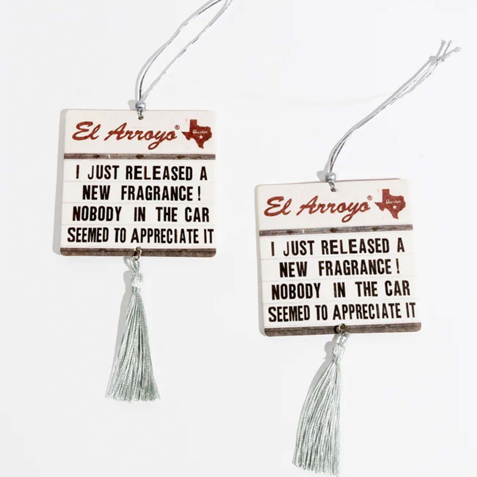El Arroyo Pack of 2 Air Fresheners, New Car Scent.  The air fresheners are in a marquee style sign, El Arroyo logo on top, the words "I just released a new fragrance! Nobody in the car seemed to appreciate it.", and a tassel hanging from the bottom.