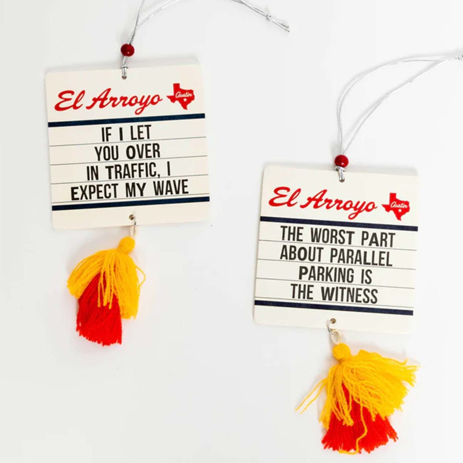El Arroyo Pack of 2 Air Fresheners, New Car Scent.  The air fresheners are in a marquee style sign, El Arroyo logo on top. First sign has the words "If I let you over in traffic, I expect my wave" The second says "The worst part about parallel parking is the witness."   There is a tassel hanging from the bottom.