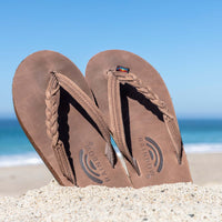 Rainbow brown braided strap flip flops displayed upright in sand with ocean backdrop.