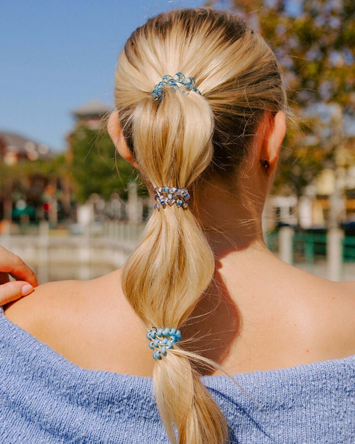 Blonde woman with low ponytail styled using multiple blue coil hair ties outdoors in natural sunlight.