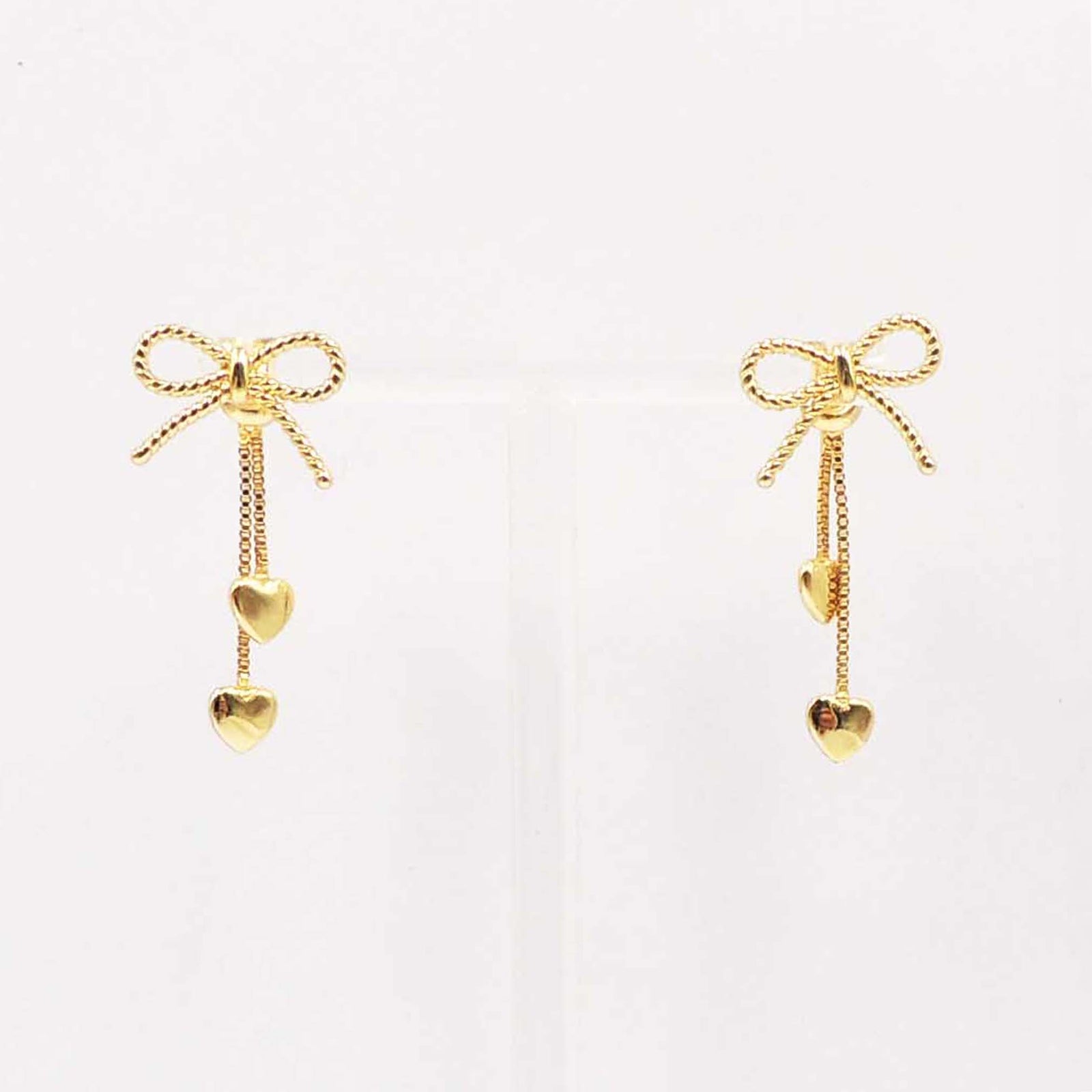 Gold-tone bow stud earrings with two dangling chain drops, each finished with small heart charms, displayed against a neutral background.