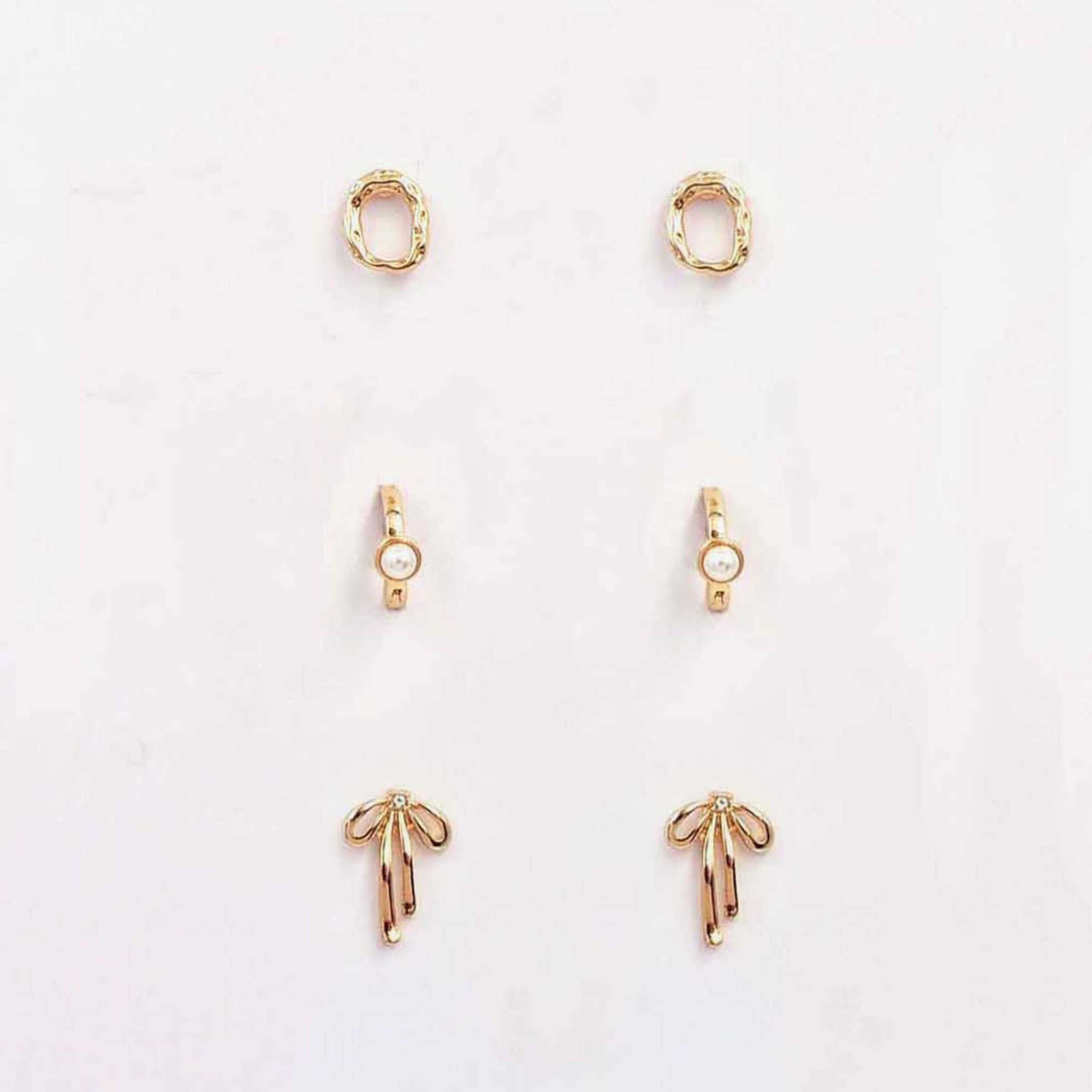 Alt text: Three-piece gold earring set featuring textured oval studs, pearl-accent huggies, and gold bow studs displayed on a card.