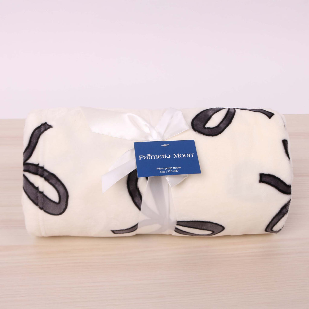 Palmetto Moon cream micro plush throw blanket rolled and tied with a white ribbon, featuring black bow pattern.