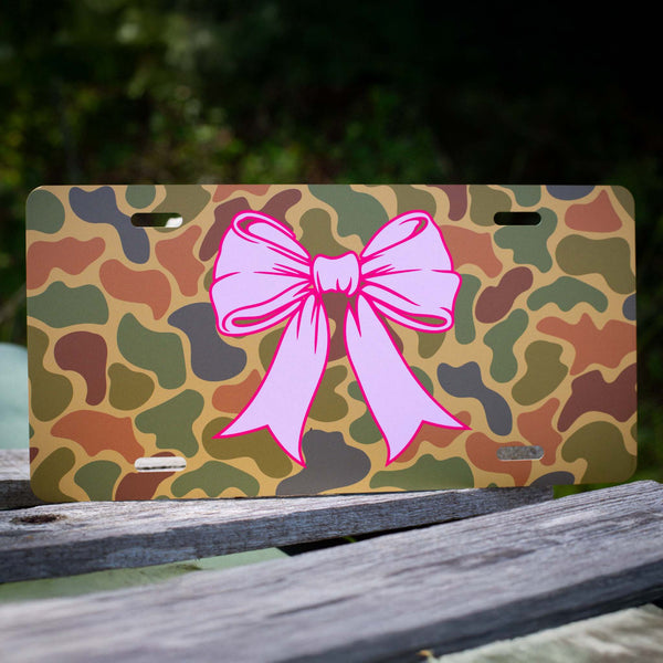 Pink bow on a camo-print aluminum license plate, displayed outdoors on a wooden surface.