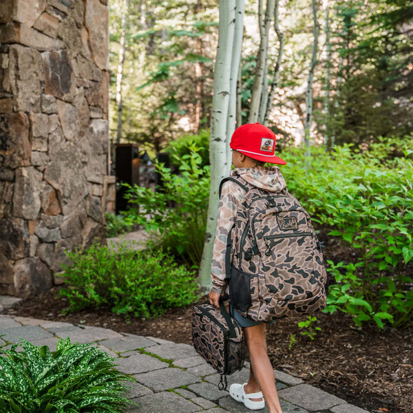 Burlebo Backpack in Guage Camo worn by a child outdoors with matching lunchbox. Backpack is large and visibly packed, shown from the back.