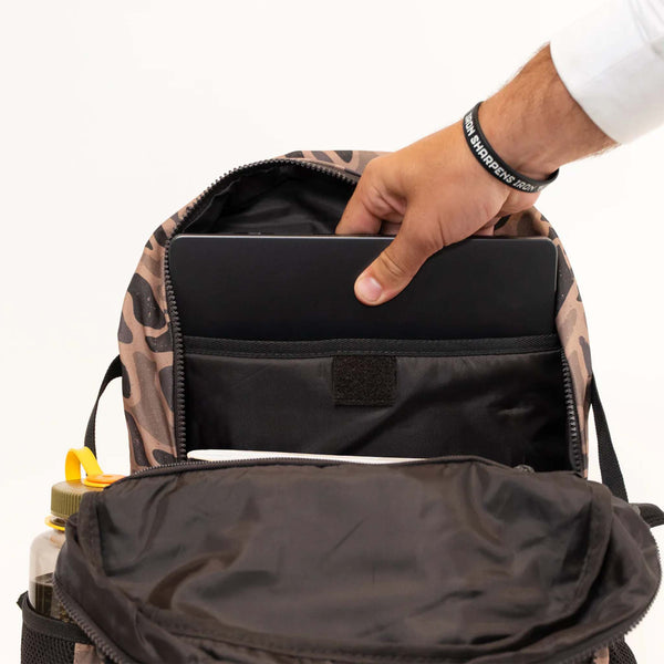 Burlebo Backpack in Guage Camo open to show a hand placing a laptop into the padded laptop sleeve in the main compartment.